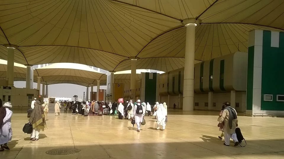 Fotografia da praça do Hajj Terminal: peregrinos a caminhar debaixo de enormes tendas bege sobre pilares brancos, à direita o edifício de fachada branca com painéis verdes